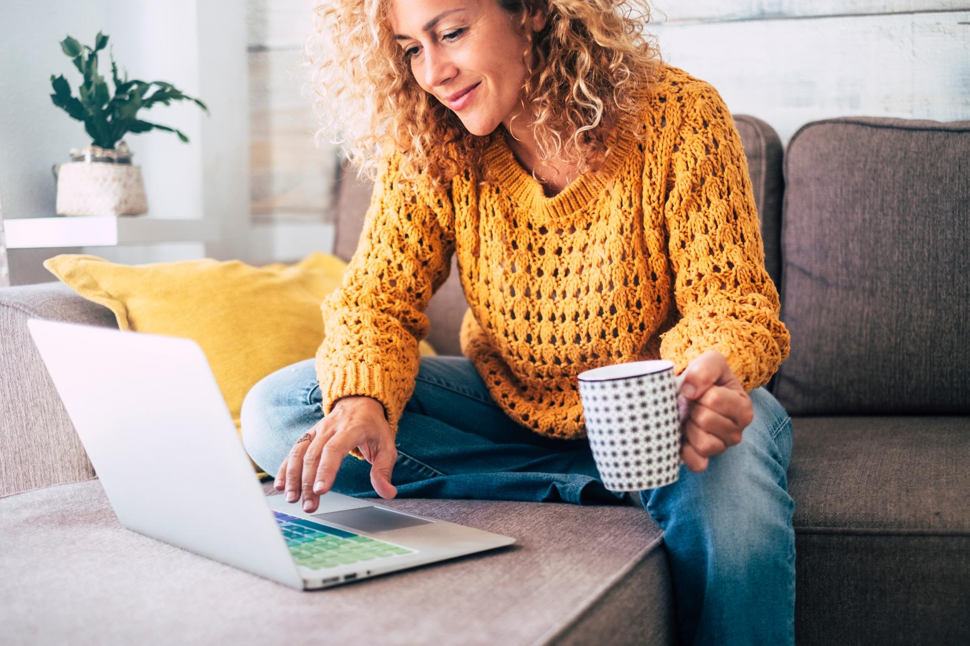 Curly-haired women is using a laptop and holding a mug in one hand