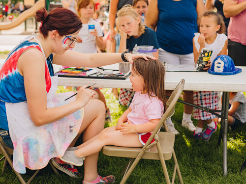 A child sitting in a folding chair gets their face painted by an artist.